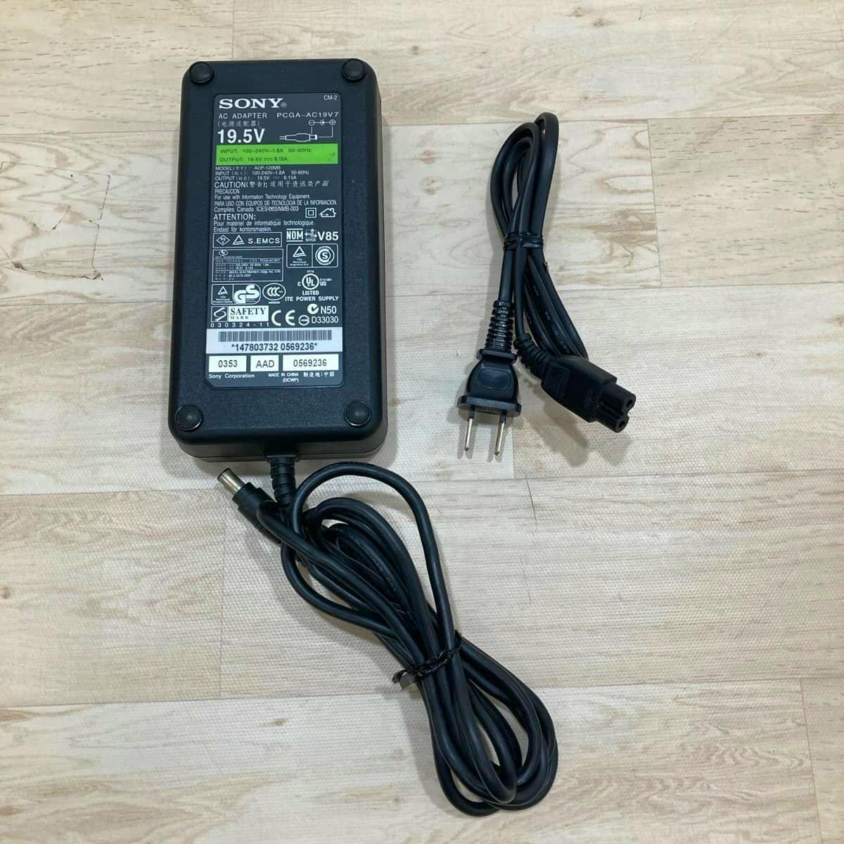 Sony black AC adapter showing the label (19.5V) with its DC output cable and a separate two‑prong AC power cord on a wooden surface.