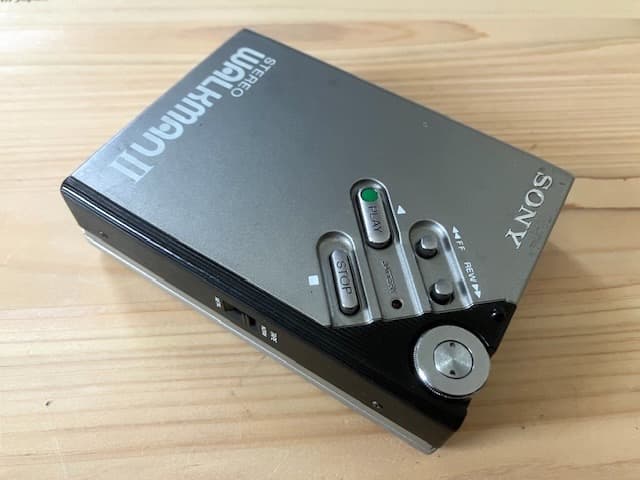 Silver Sony Walkman II (WM-2) cassette player on a wooden surface showing control buttons and a rewind wheel.