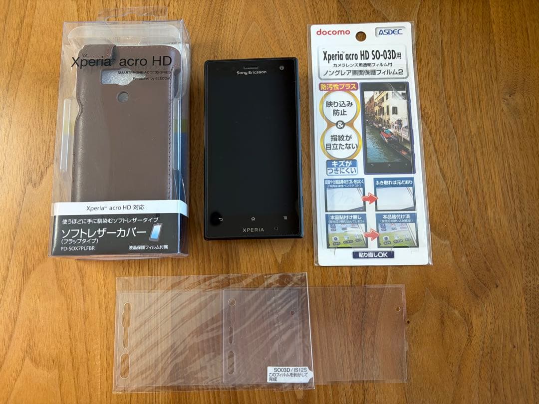 Black Sony Xperia SO-03D smartphone shown with a packaged cover and screen protectors on a wooden surface.