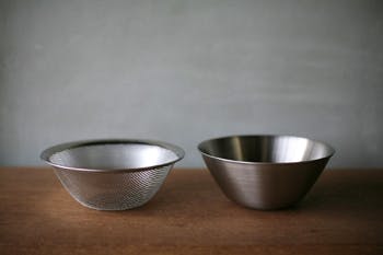 Two stainless steel bowls, one solid and one mesh strainer, shown on a wooden surface with a gray background.