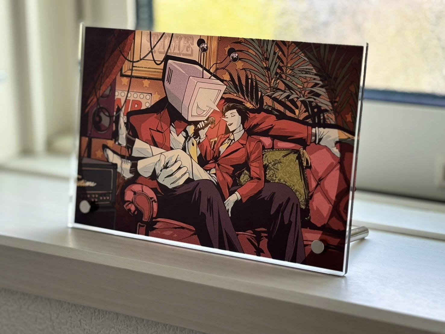 An acrylic photo panel standing on a windowsill displaying printed artwork of two characters on a red sofa, mounted with two round standoffs and a metal support.