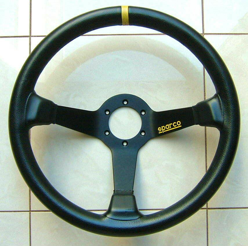 Black SPARCO leather deep-cone three-spoke steering wheel shown flat on a tiled floor with a yellow top-center stripe and Sparco logo.