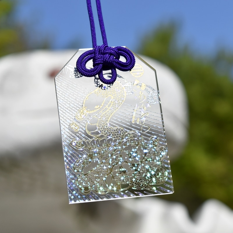 Photograph of the amulet showing a glittering white-snake design on a reflective tag hanging from a purple cord.
