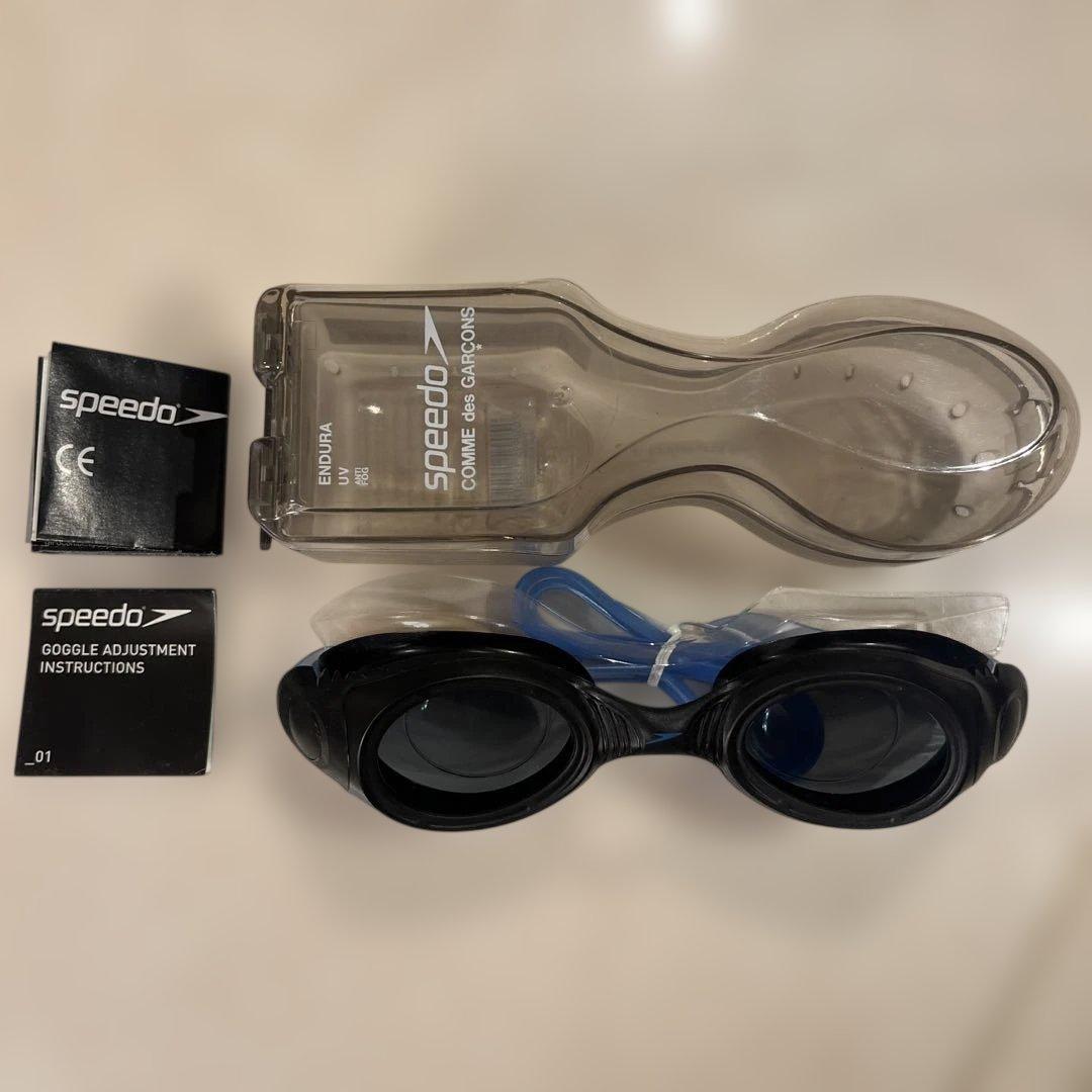 A pair of black and blue Speedo × GARÇONS swimming goggles with a clear plastic case and Speedo instruction booklets on a light surface.