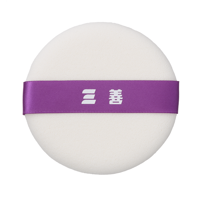 Round white powder pad sponge puff with a purple ribbon strap across the top bearing white Japanese characters.