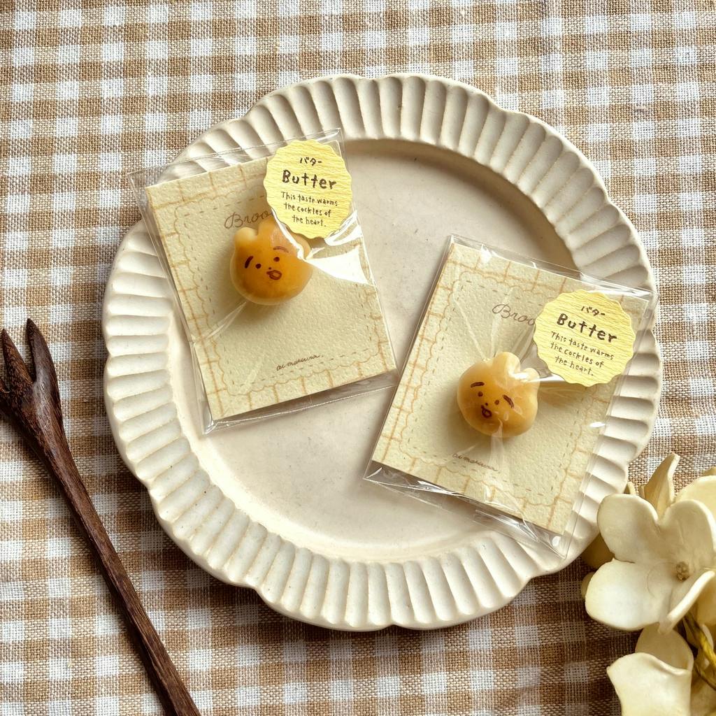 Two packaged pins (squirrel-shaped bread-face brooches) displayed on a ceramic plate over a beige gingham cloth.