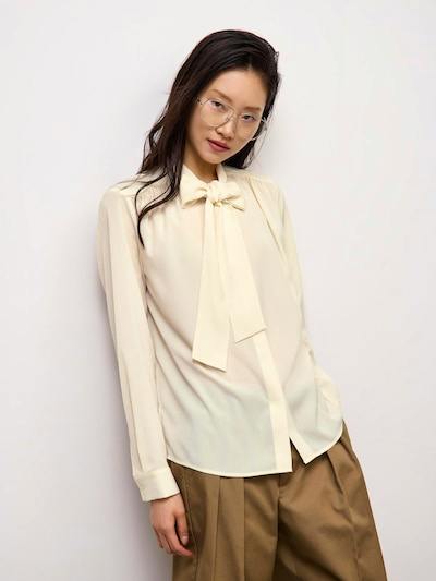 Model wearing a cream stand-collar blouse with a bow tie at the neck, shown tucked over brown trousers.