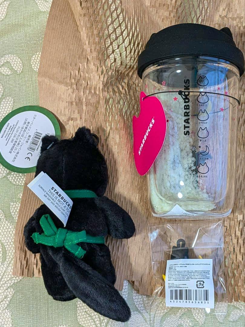 A Starbucks Halloween set showing a heat-resistant glass cup with a black silicone lid and visible Starbucks logo, a black Bearista plush wearing a green apron, and a small packaged cap figure.