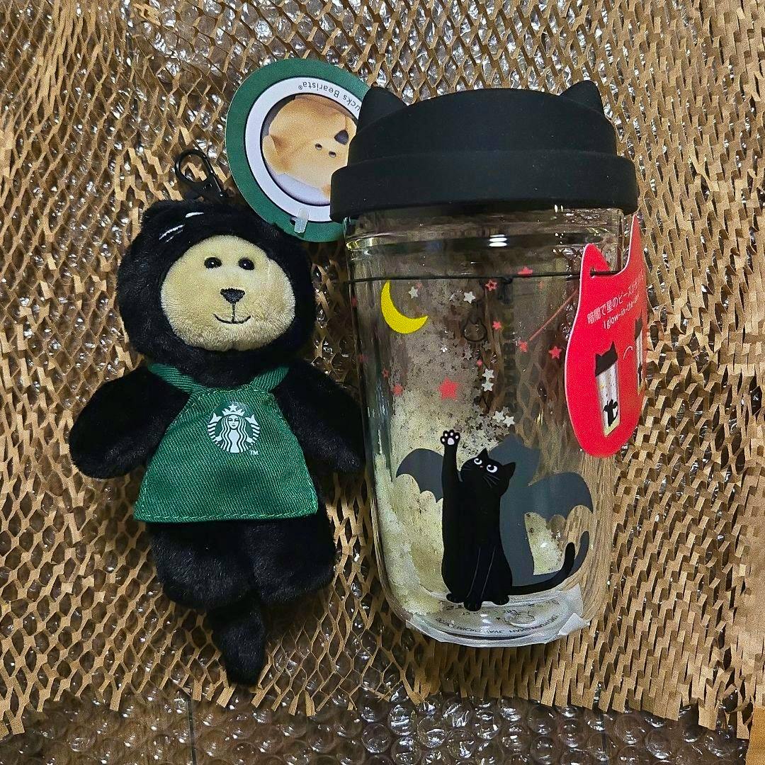 A Starbucks Halloween Bearista plush keychain wearing a green apron next to a heat-resistant glass mug with a black cat and moon design and a black silicone lid.