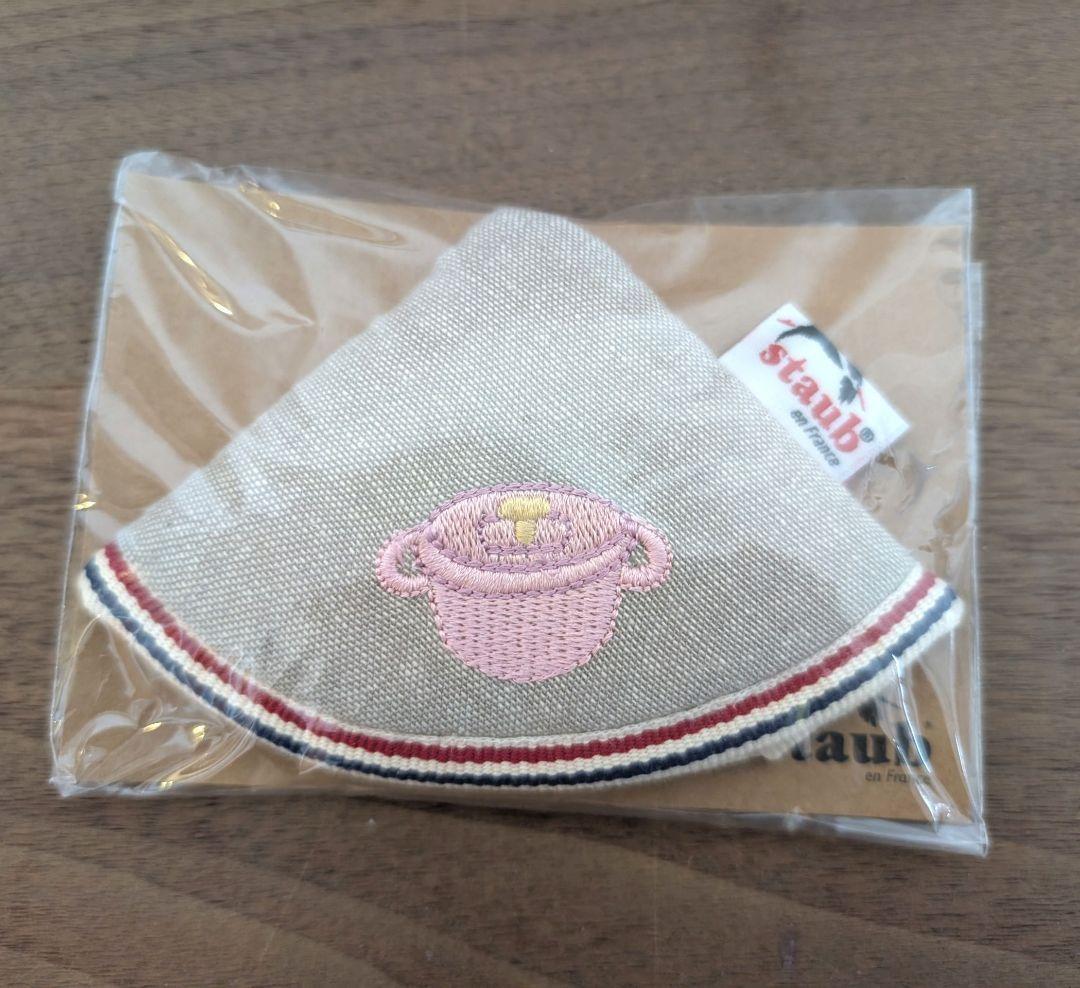 Staub cotton pot holder in sorbet rose shown in plastic packaging with an embroidered pink pot, on a wooden table.