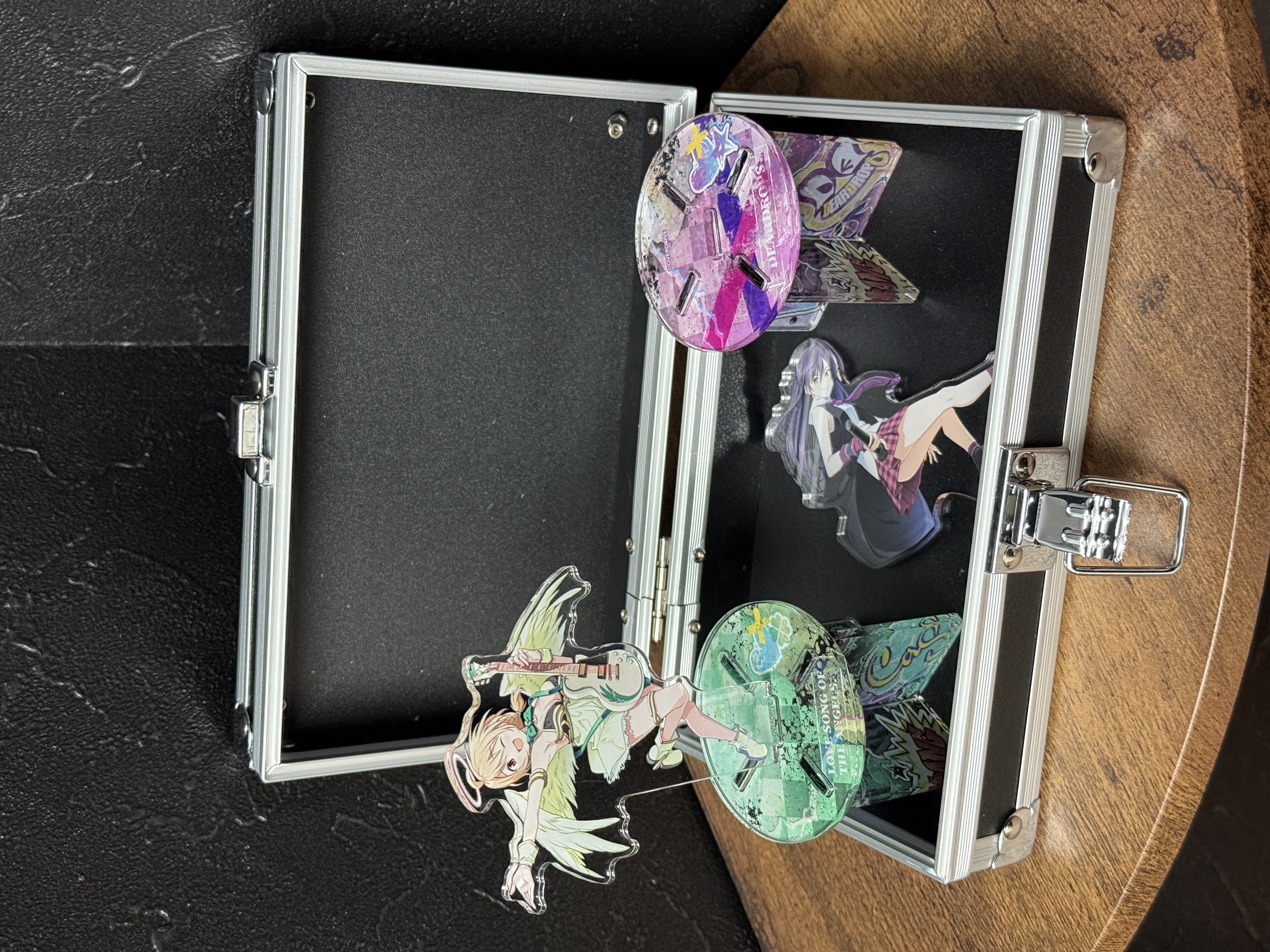 Open steel attache case (Attache Case Multi Box) with black interior on a wooden surface, holding small acrylic figure stands.