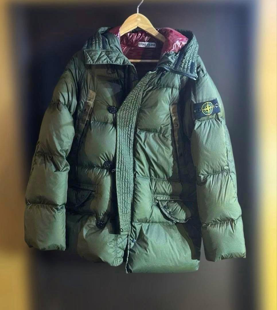 Khaki Stone Island men's down jacket (polyester) hanging on a wooden hanger against a dark background.