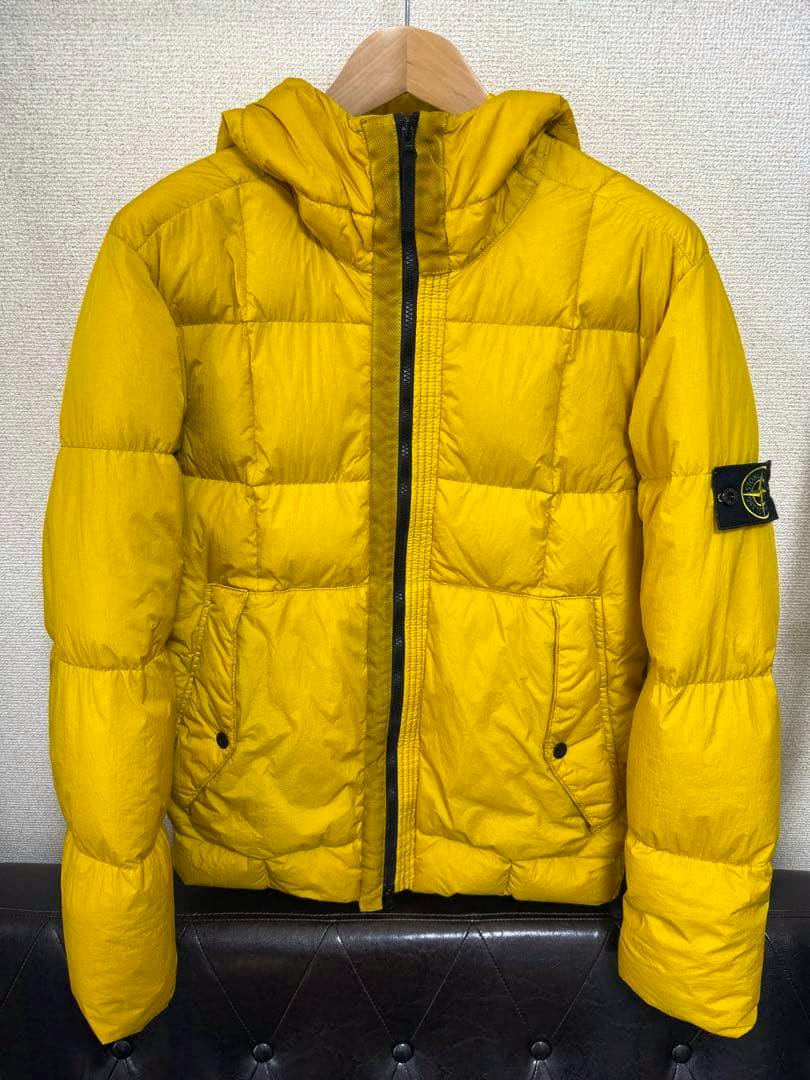 Front view of a yellow men's nylon down jacket with hood, black center zipper, front pockets, and a patch on the left sleeve hanging on a wooden hanger.