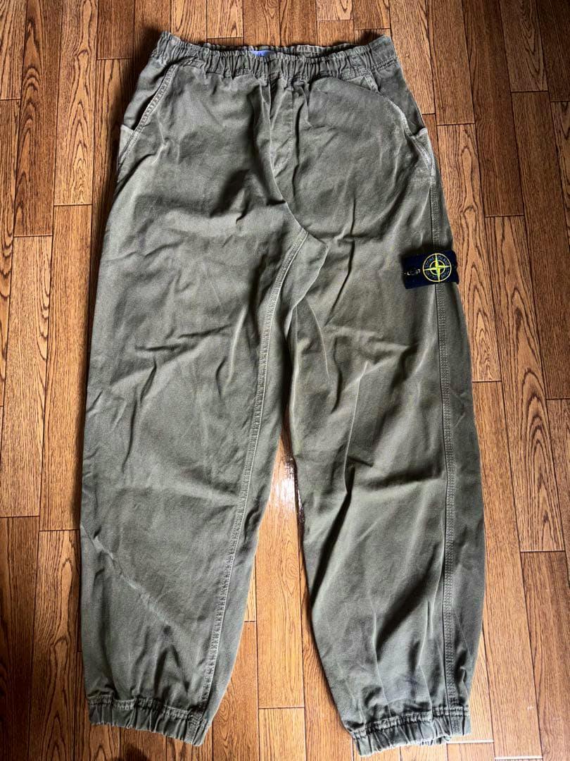 Olive-green Stone Island pants laid flat on a wooden floor showing the full front view with the brand badge on the leg.