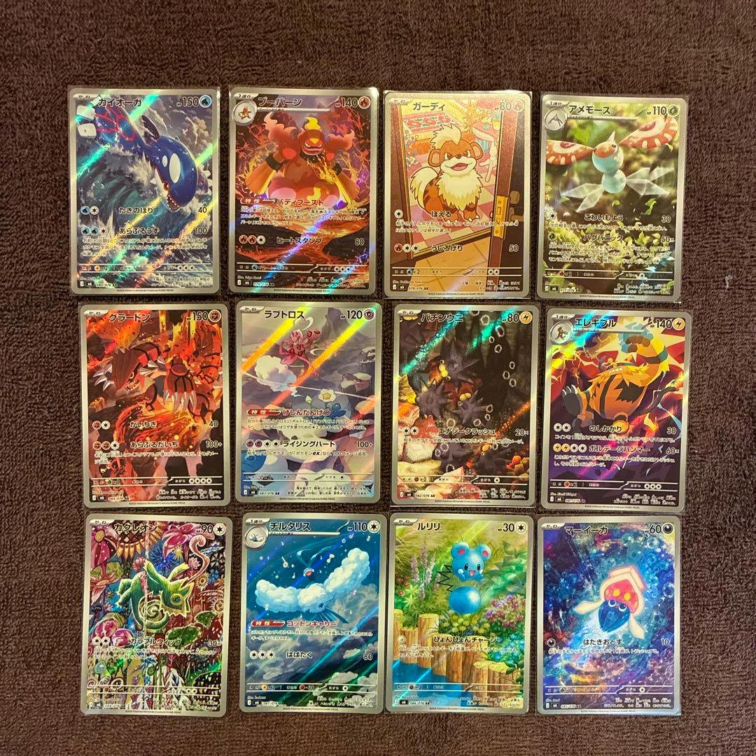 Twelve Japanese holographic trading cards arranged in a 4x3 grid on brown fabric, photographed from above.