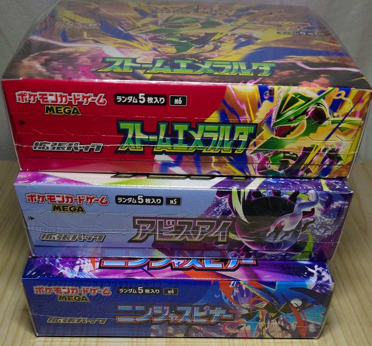 Three shrink-wrapped Pokémon booster boxes (Storm Emeralda top, Abyss Eye middle, Ninja Spinner bottom) stacked on a wooden surface.
