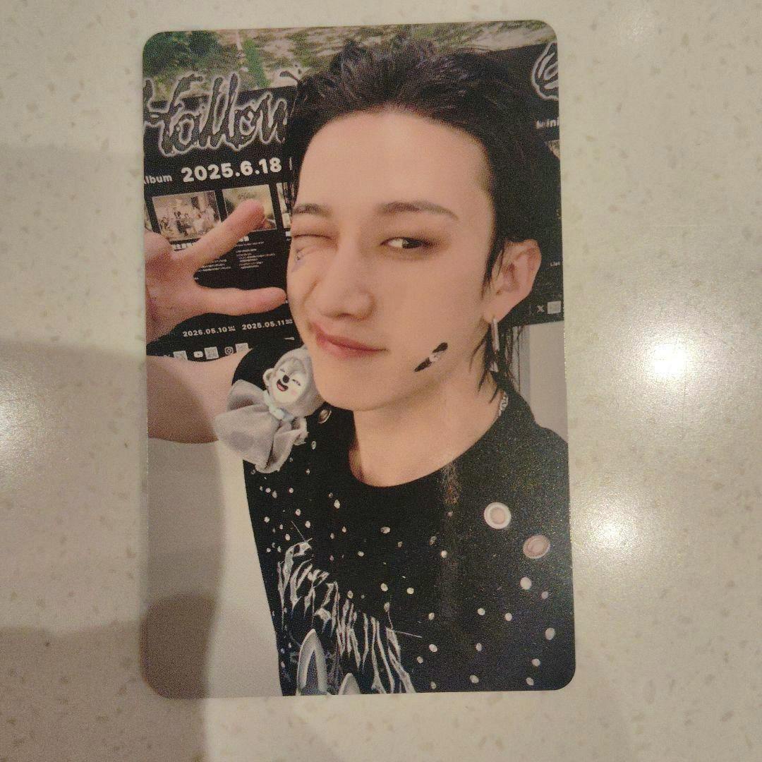 Photocard showing a winking person making a peace sign, wearing a patterned black shirt and a small plush accessory on the shoulder.