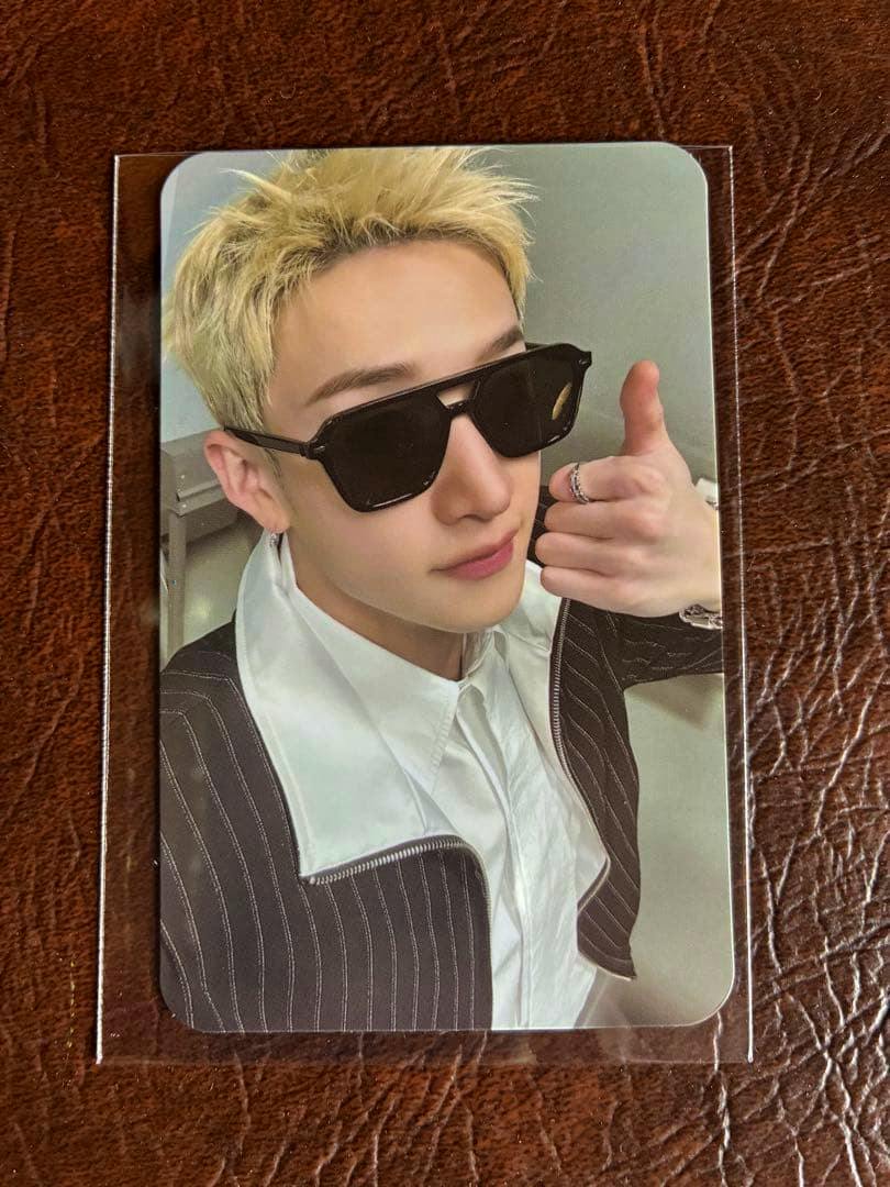 Photocard in a clear sleeve showing a blonde person wearing sunglasses and a pinstripe jacket on a brown leather surface.