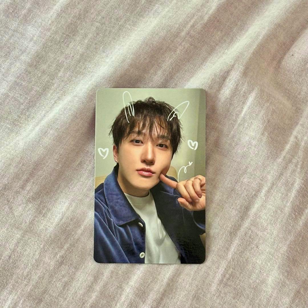 A photocard showing a portrait of a young man wearing a blue jacket with white doodle ears and hearts, placed on a pale fabric background.