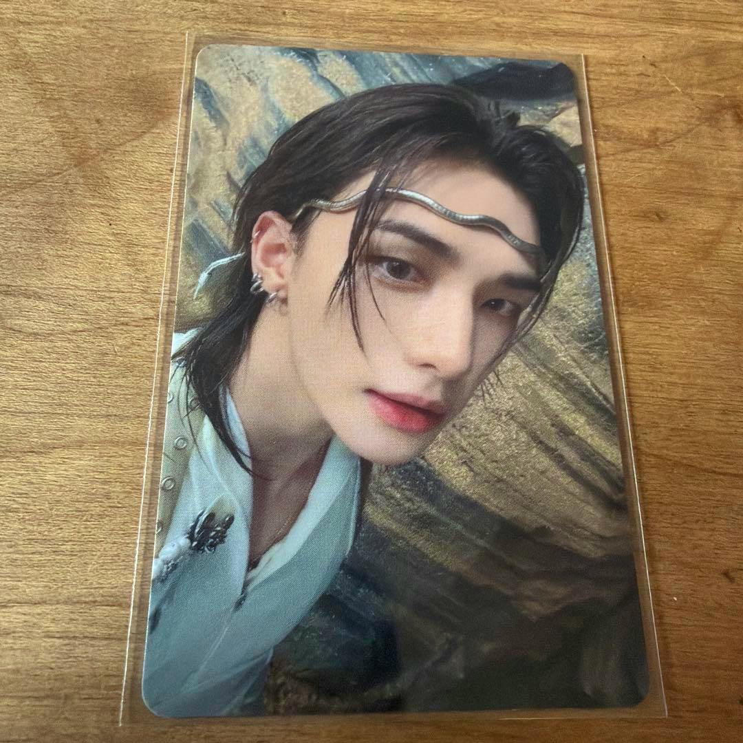Photocard in a clear sleeve showing a close-up portrait of a young person with dark hair and a decorative headband, placed on a wooden surface.