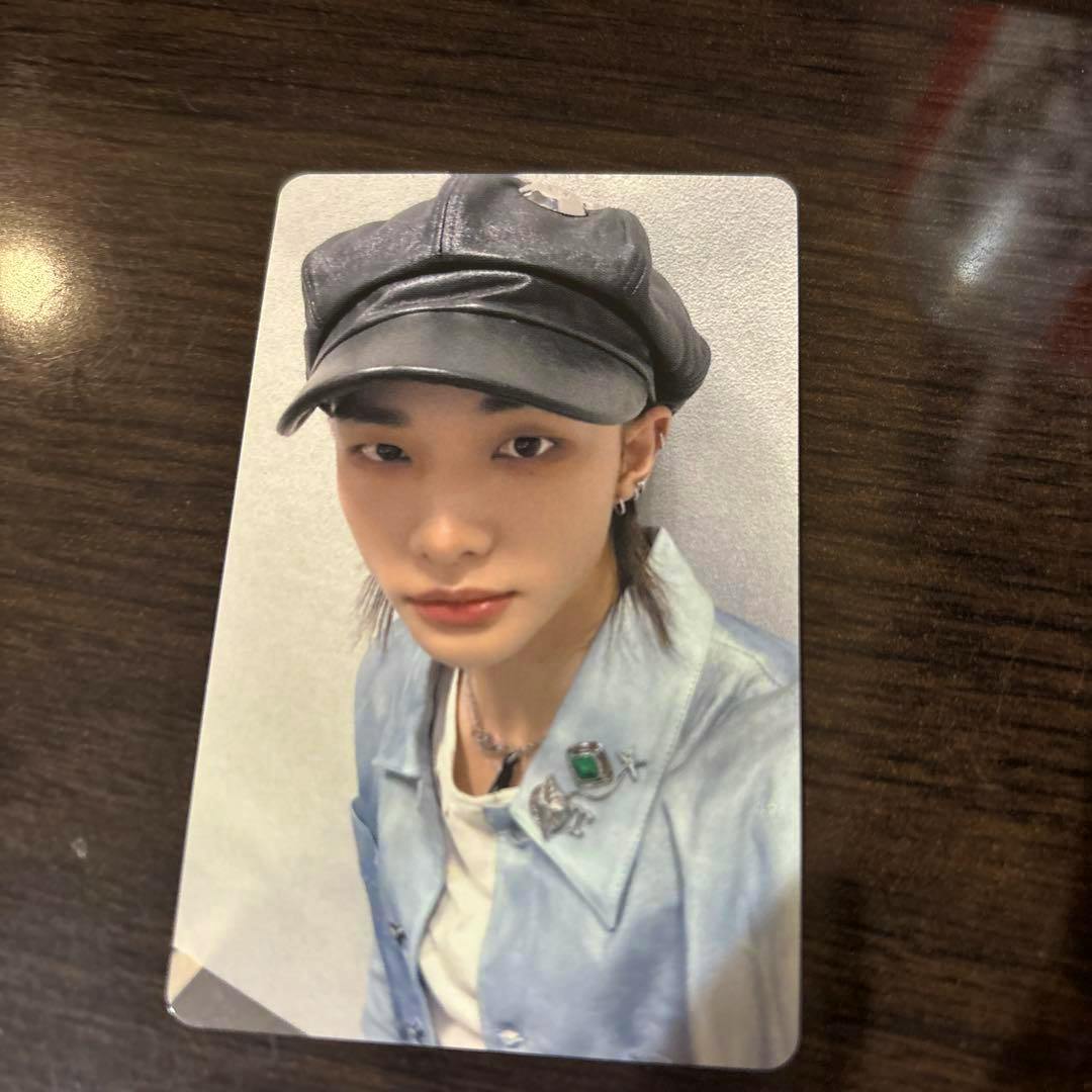 Photocard of Stray Kids member Hyunjin showing a close-up portrait of him wearing a black cap and light-blue jacket on a dark wood surface.