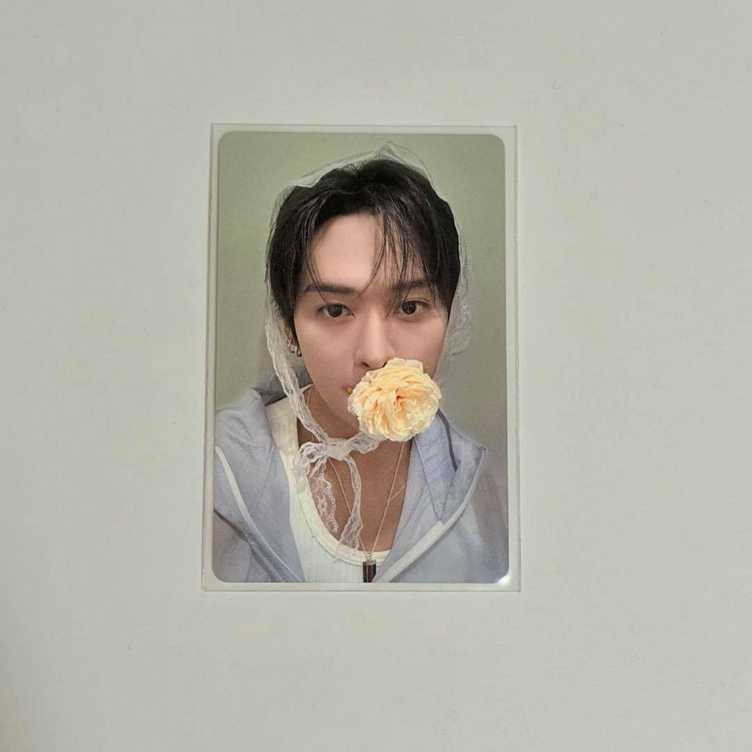 Photocard showing a close-up portrait of a person wearing a light jacket and lace headscarf holding a pale yellow flower to their mouth on a white background.