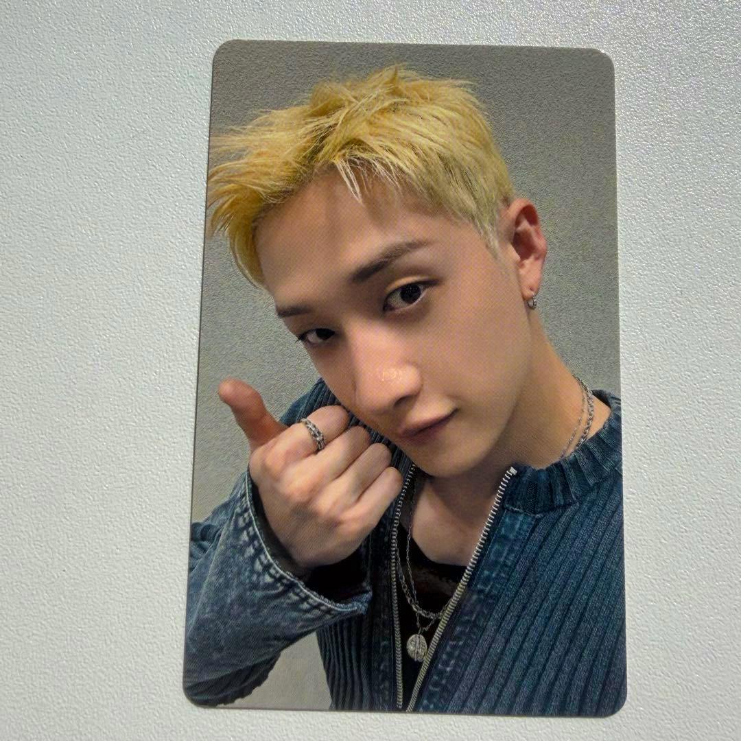 Photocard showing a close-up selfie of a blond-haired person wearing layered necklaces and a zip-up top.