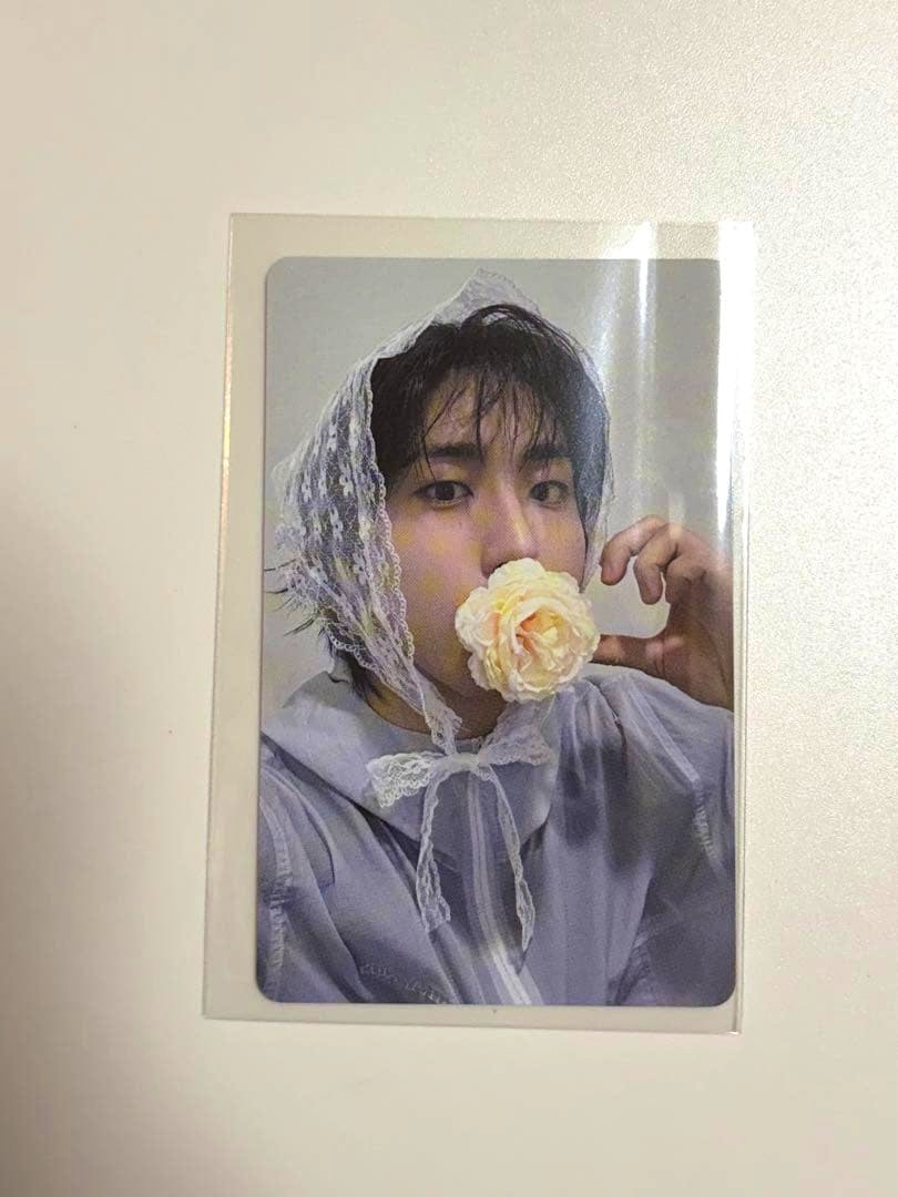 Front of a photocard in a clear sleeve showing a portrait of a person wearing a lace headscarf and holding a flower to their mouth.