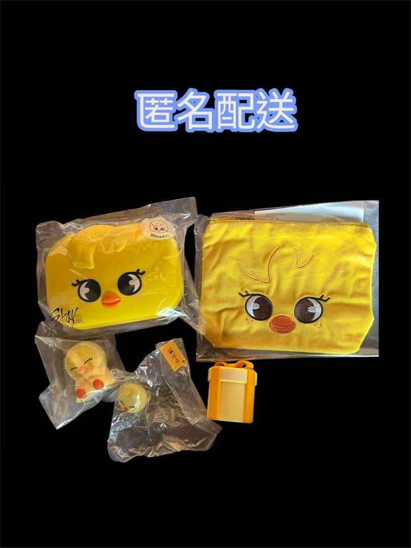 Photograph of the Stray Kids SKZOO BbokAri four-piece set showing a yellow pouch, a yellow mini box, small character figures, and a small accessory arranged on a black background.