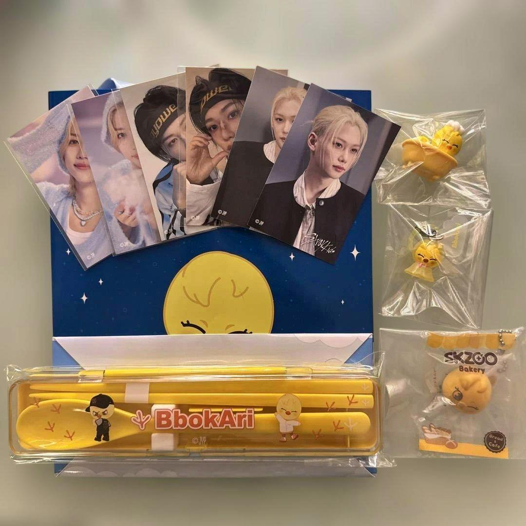 A flat lay of Stray Kids SKZOO BbokAri goods showing member photocards, a yellow BbokAri cutlery set in a case, and several packaged charms.