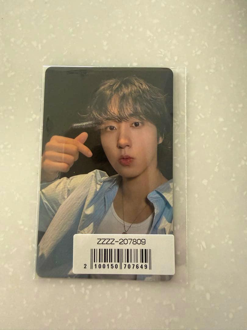 A photocard in a clear plastic sleeve shows a portrait of a person making a finger-heart gesture, with a barcode sticker on the lower front.