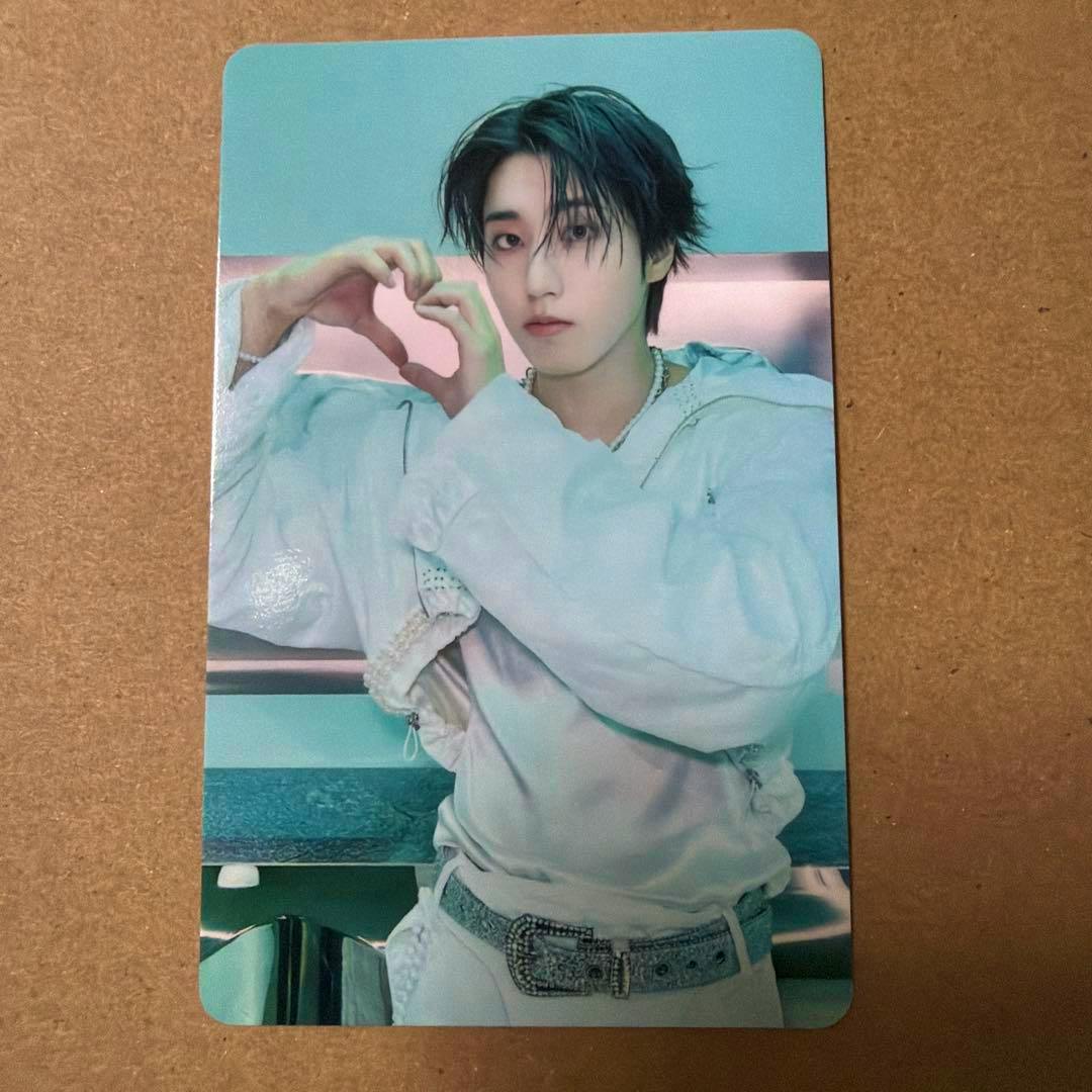 A photocard showing a person wearing a light jacket making a heart shape with their hands against a teal background.