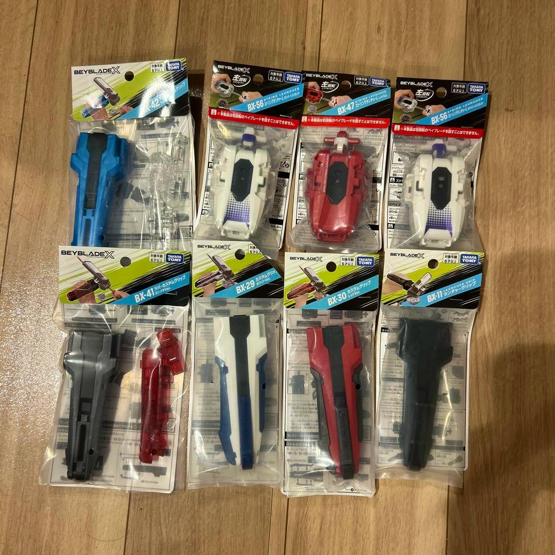 Multiple packaged string launchers and custom grips for Beyblade toys arranged on a wooden floor.