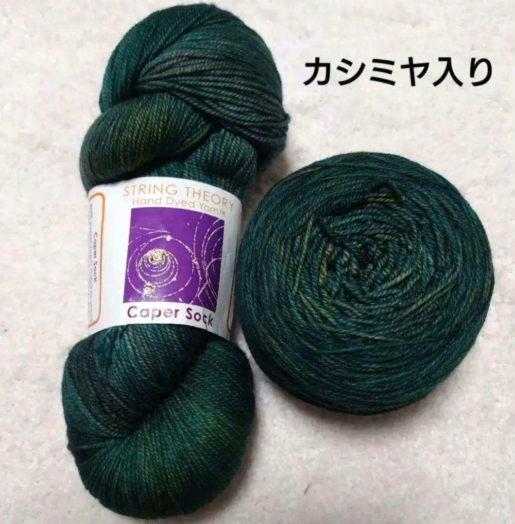 Two dark green hand-dyed String Theory sock yarn skeins (cashmere blend), one hank and one cake with label visible.