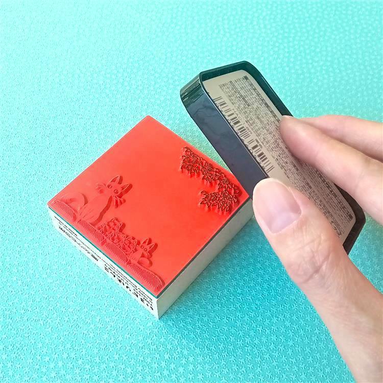 A wooden Studio Ghibli “Laputa” stamp showing the red rubber stamp surface next to a hand holding an ink pad on a turquoise background.