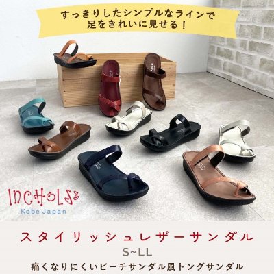 Photo of multiple leather sandals in various colors arranged on a floor with a wooden block backdrop.