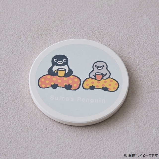 Round absorbent cork coaster showing two Suica penguins holding cups on a pale background.