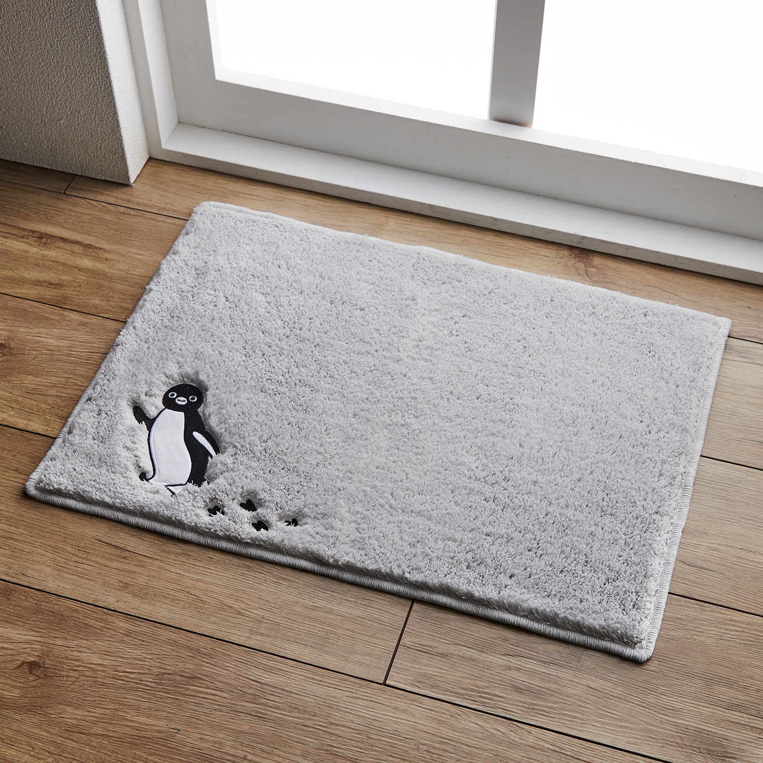 Gray bath mat with a Suica penguin appliqué and paw prints placed on a wooden floor near a window.