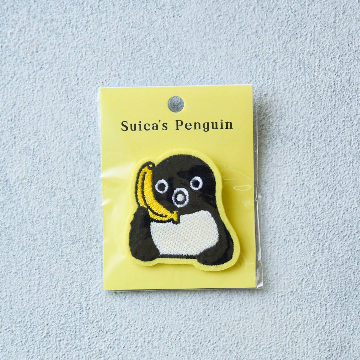 Embroidered Suica Penguin pin badge holding a banana on a yellow backing card in clear plastic packaging.