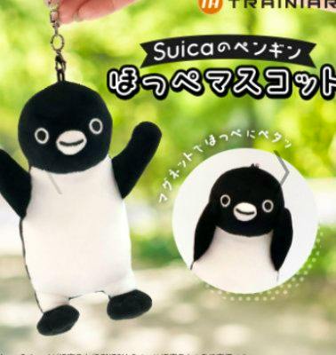 Plush Suica penguin keychain (ほっぺマスコット) held by a hand against a blurred green background.
