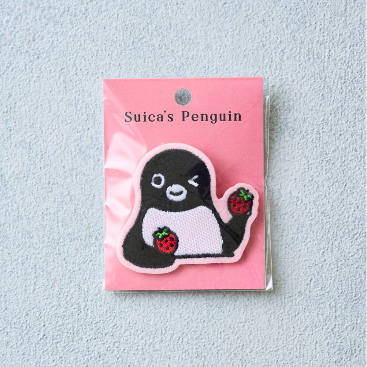 A Suica's Penguin sagra embroidered pin badge with strawberries mounted on a pink backing card inside clear plastic packaging.