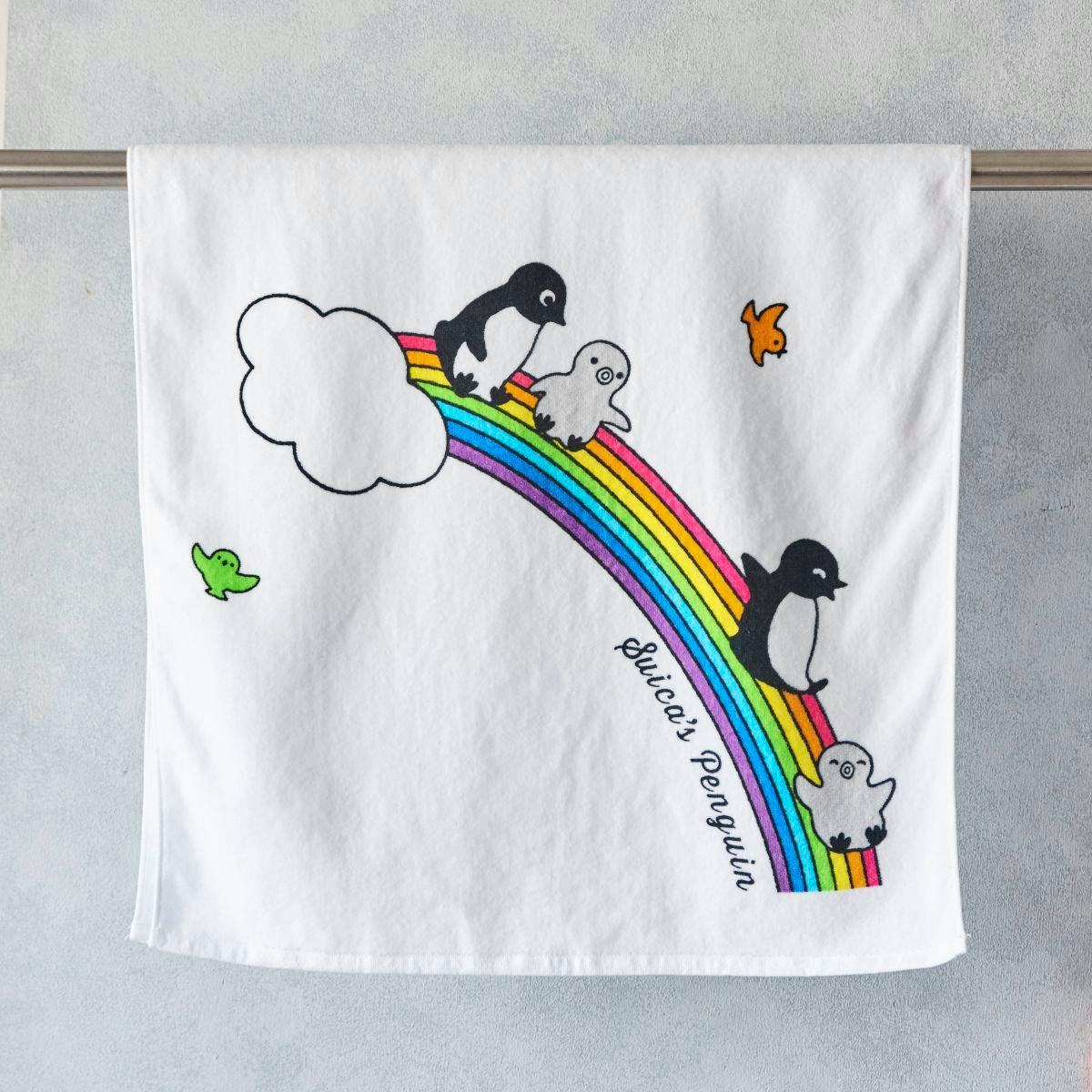 White cotton bath towel showing Suica's Penguin characters on a rainbow design, hung over a metal rod.