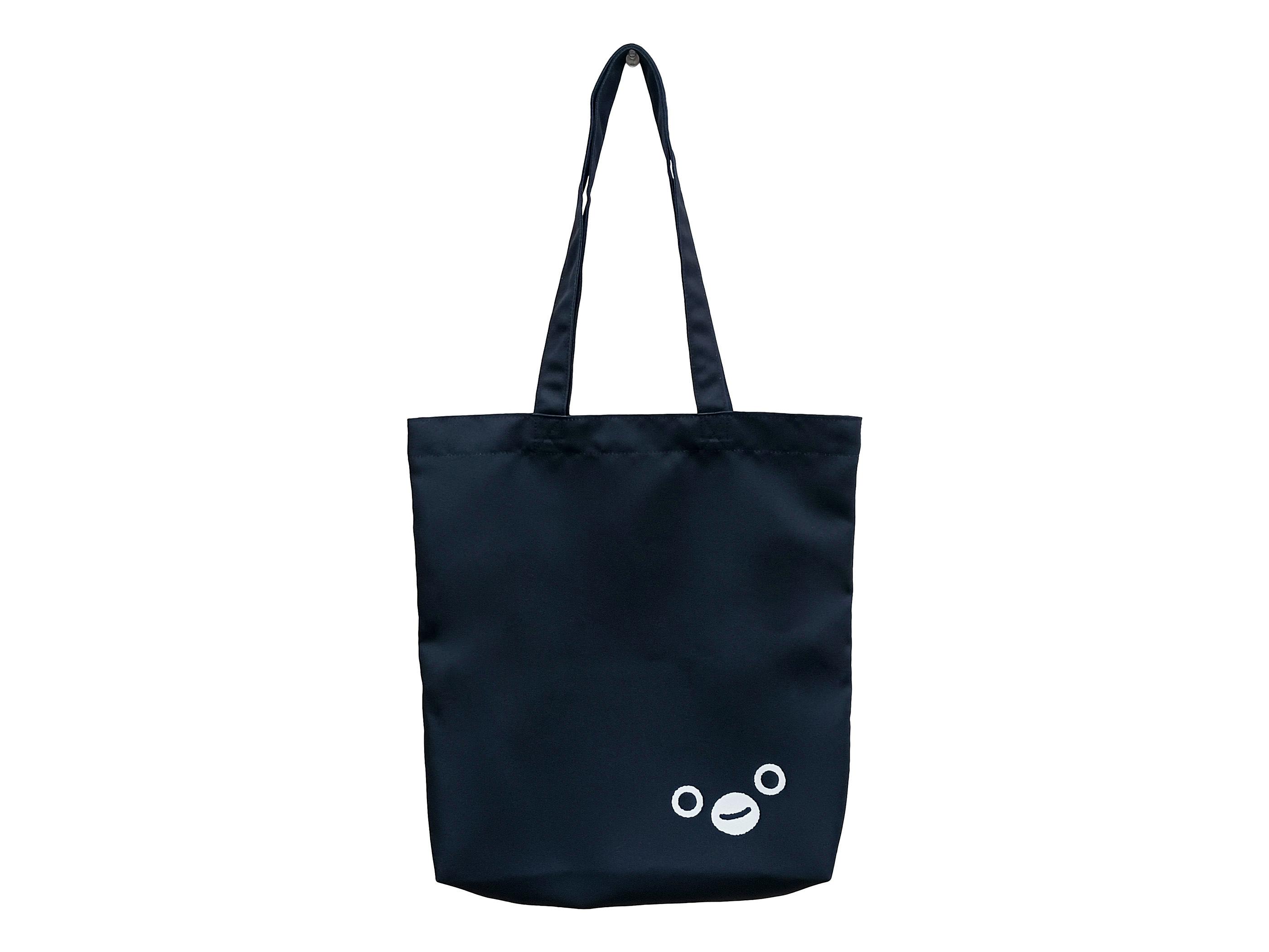 Black tote bag with long shoulder straps and a small white Suica penguin face printed near the lower right on a white background.