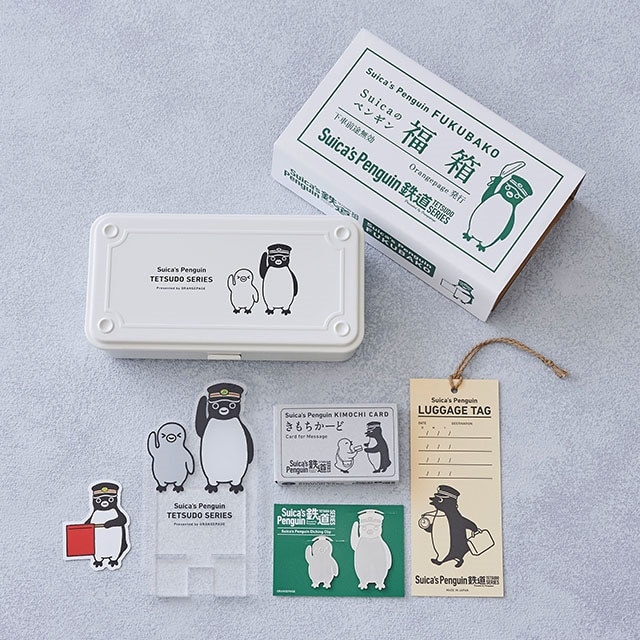 Closed white metal Suica Penguin tool box with penguin illustration, shown with its outer box, stickers, and a luggage tag arranged around it.