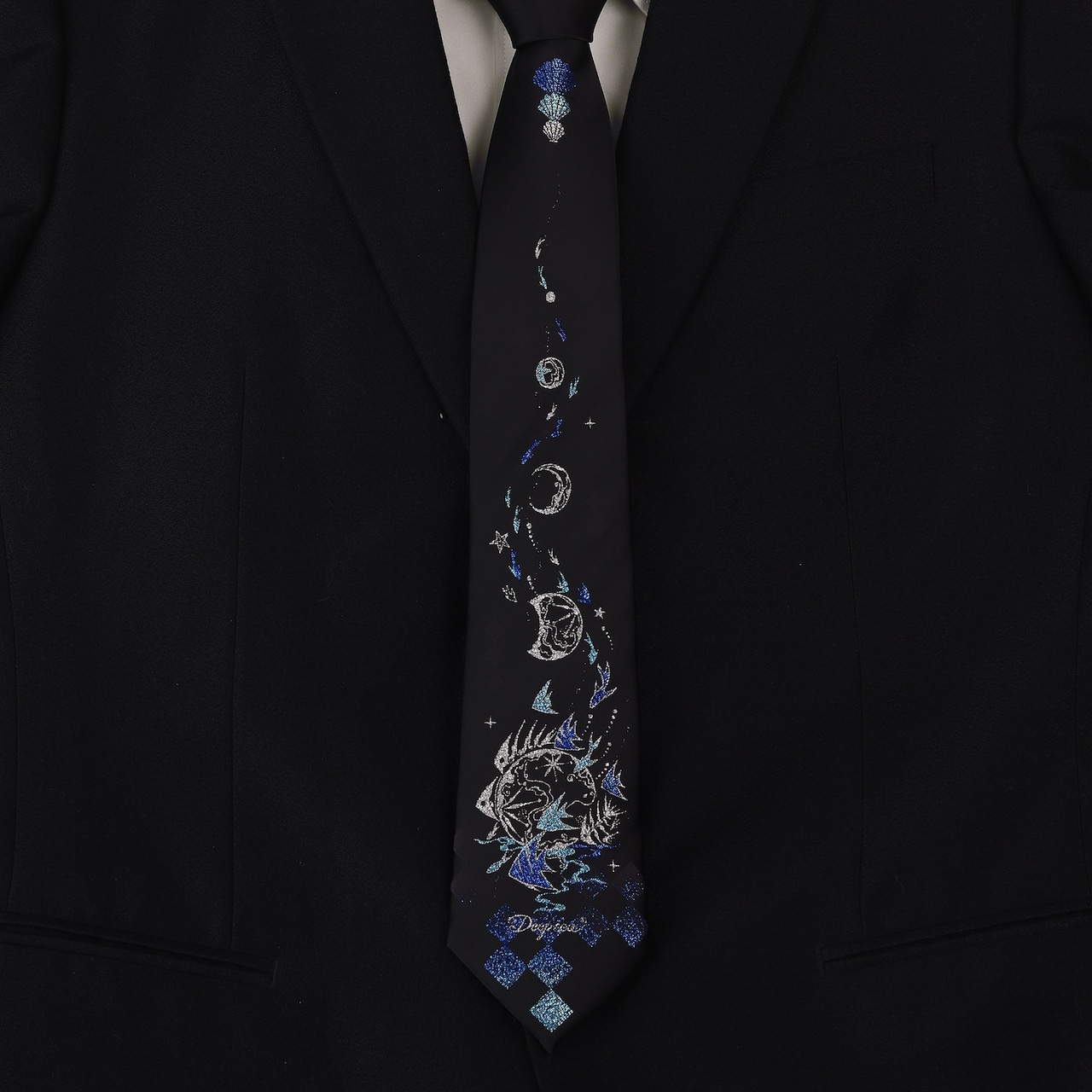 Black polyester necktie with a vertical blue and silver moon, stars, and checkerboard pattern shown knotted on a black suit jacket.