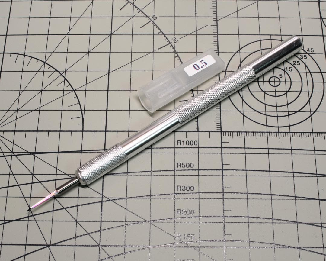 Silver chisel (0.5 mm) with a textured metal handle and a clear plastic cap labeled “0.5” lying on a gridded cutting mat.