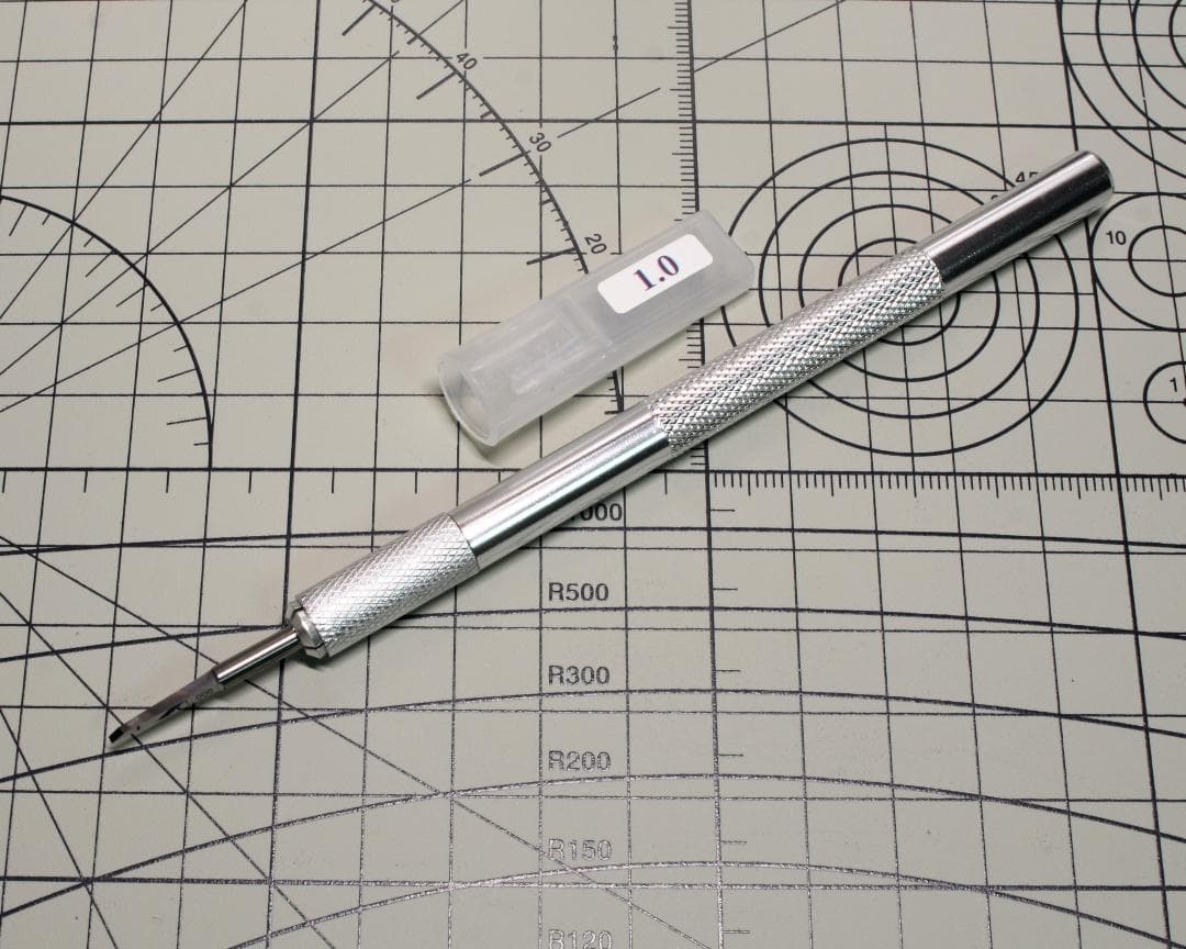 A silver knurled-handled chisel with its blade and a plastic cap labeled "1.0" placed on a gridded cutting mat.