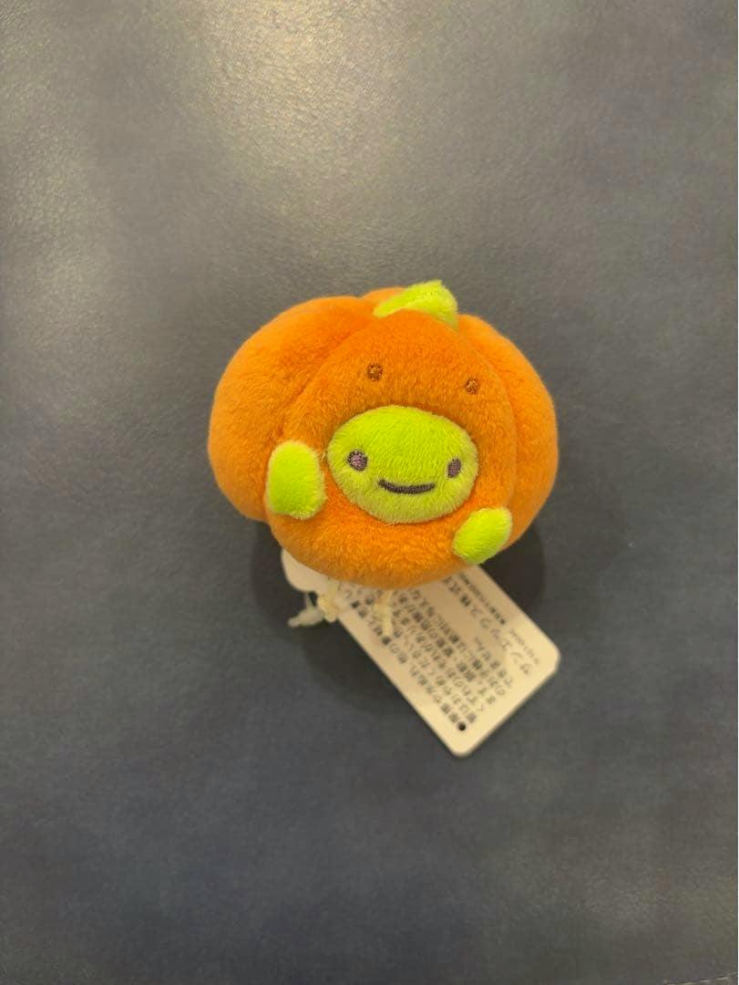 Orange pumpkin-themed Sumikko Gurashi てのりぬいぐるみ (ざっそう) palm-sized plush with its tag on a dark surface.