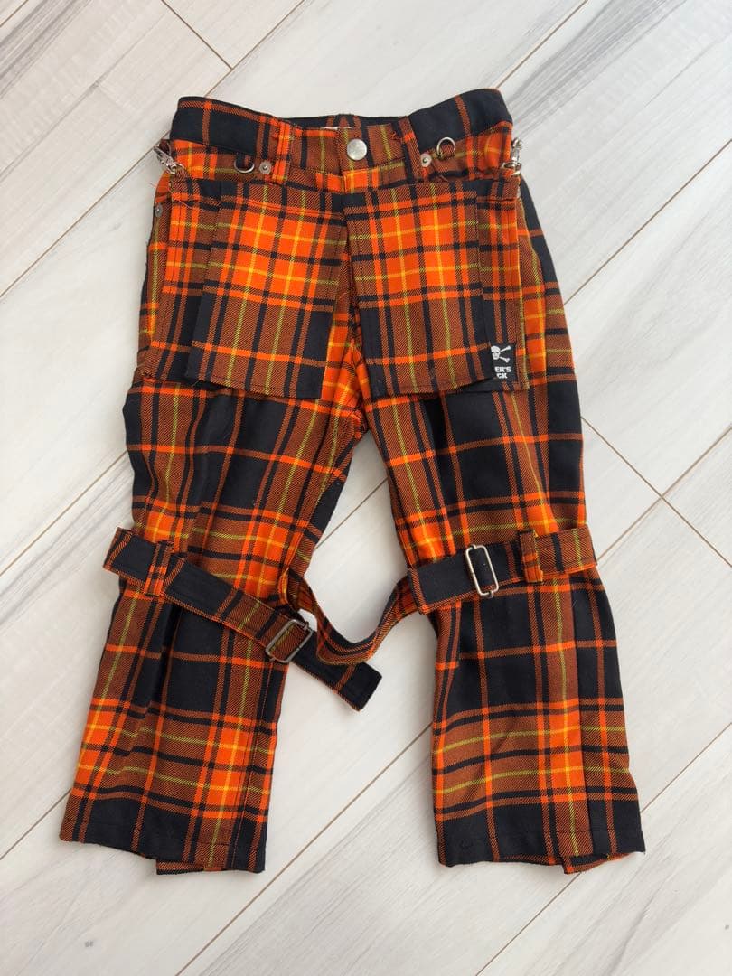 Orange and black plaid women's bondage-style cotton pants with straps and metal hardware laid flat on a light wood floor.