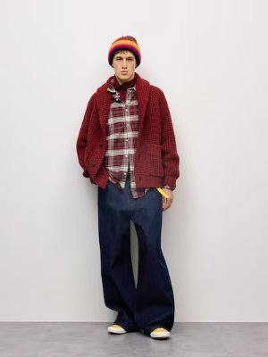 A model wearing dark super-wide denim jeans paired with a red knit cardigan and a beanie, standing against a white background.